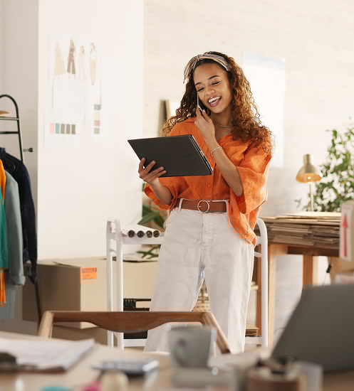 A smiling woman wearing an orange blouse and white pants stands in a bright office space, holding a tablet and talking, possibly during a video call or presentation.